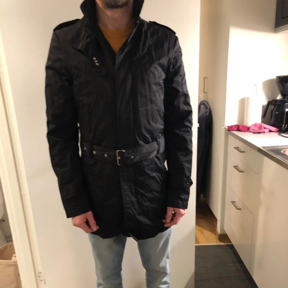 Säljer en stilren svart trenchcoat från J.Lindeberg. Jackan har ett bälte i midjan och långa ärmar. Perfekt för höst och vår med sin blandning av bomull och nylon. Den har knappar och en diskret design som passar till många tillfällen.. Takit.