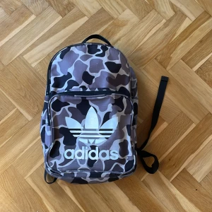 Camouflage ryggsäck från Adidas - Snygg ryggsäck från Adidas med ett coolt camouflage-mönster i svart, grått och beige. Väskan har ett stort huvudfack och en praktisk framficka med dragkedja. Perfekt för skolan eller träningen! Det finns några fläckar på själva trycket Adidas