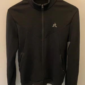 Svart zip up från Le Coq Sportif - Säljer en stilren svart zip up från Le Coq Sportif med dragkedja och hög krage. Jackan har en diskret logga på bröstet och praktiska fickor med dragkedja. Perfekt när som helst. Fungerar på sommar kvällarna men även på hösten och våren, funkar också bra för uppvärmnings kläder eller bara vardagligt.