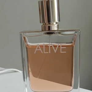 80 ml Parfym 'Alive' från Boss - Elegant parfymflaska med namnet 'Alive' från Boss. Flaskan är rektangulär med en stilren design och har en gyllene kork. Innehållet är en varm, bärnstensfärgad vätska som ger en lyxig känsla.