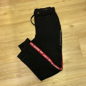 Svarta mjukisbyxor med röd detalj - Snygga svarta mjukisbyxor med en cool röd remsa längs sidan där det står 'REMOVE BEFORE FLIGHT'. Byxorna har en dragkedja på benet och snörning i midjan för bästa passform. Perfekta för en avslappnad stil.