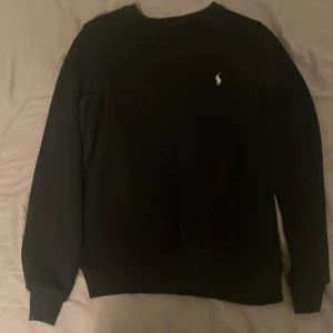 Svart tröja från Ralph Lauren - Säljer en stilren svart tröja från Ralph Lauren med det klassiska logotypen broderad i vitt på bröstet. Tröjan har långa ärmar och en rund halsringning. Perfekt för en avslappnad och snygg look.