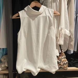Vit ärmlös blus från H&M - Säljer en stilren vit ärmlös blus från H&M. Blusen har en klassisk krage och en diskret dragkedja i ryggen för en elegant look. Perfekt för både vardag och speciella tillfällen.