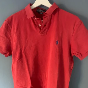 Röd pikétröja från Ralph Lauren - Snygg röd pikétröja från Ralph Lauren med klassisk krage och knappar framtill. Den har en broderad logga i blått på bröstet. Perfekt för en stilren look.