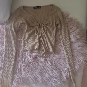 Beige ribbad topp från F3 - Säljer en beige ribbad topp från F3 i storlek XS. Toppen har långa ärmar och en knytning framtill som ger en snygg detalj. Perfekt för en stilren look.