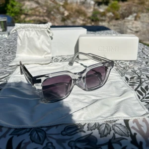 Grå solglasögon från Chimi - Snygga grå solglasögon från Chimi med transparent båge och mörka linser. Perfekta för soliga dagar och kommer med ett stilrent fodral. Modellen är 04M Grey.
