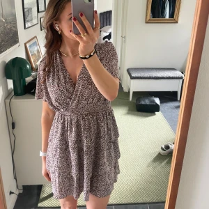 Blommig klänning från chealse  - Säljer en söt blommig klänning från Vero Moda i storlek L men skulle säga att den är mer som en M. Klänningen har en omlott-design med korta ärmar och volangdetaljer. Aldrig använd