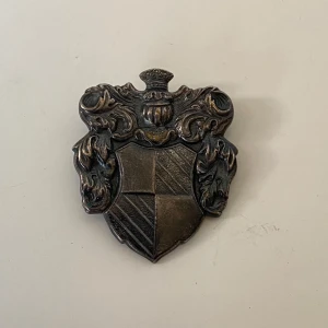 Vintage brosch i form av ett vapensköld - Säljer en vintage brosch i form av en detaljerad vapensköld. Broschen har en mörk silverfärg och är dekorerad med intrikata mönster och en krona på toppen. Perfekt för att ge en historisk touch till din outfit.