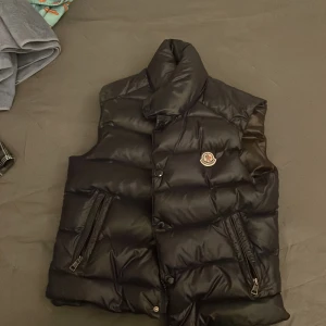 Svart dunväst från Moncler -  ÄKTA )Snygg svart dunväst från Moncler med dragkedja och knappar. Perfekt för kyliga dagar med sin quiltade design och stilrena look. Västen har en klassisk Moncler-logga på bröstet. Den är i storlek 0 som motsvarar xs- s men jag kan fortfarande använda den och jag använder medium i kläder! Kan skicka mer bilder om det önskas. Västen är i toppen skick 