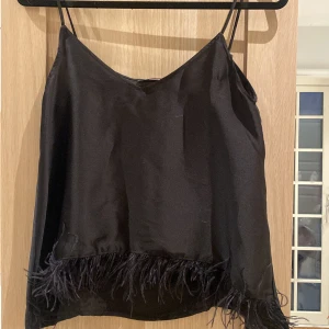 Svart linnetopp från Zara med fjädrar - Snygg svart linnetopp från Zara med tunna axelband och dekorativa fjädrar längs nederkanten. Perfekt för en stilren och trendig look. Tillverkad i ett glansigt material som ger en lyxig känsla.