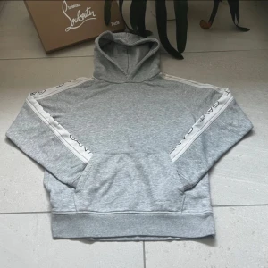 Grå hoodie från GANT - Säljer en stilren grå hoodie från GANT med vita detaljer och logga längs ärmarna. Tröjan har en klassisk känguruficka och en bekväm huva. Perfekt för en avslappnad stil.