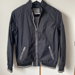 Fin vårjacka junior från DNMZ - Stilren svart vindjacka från DNMZ med dragkedja, ribbade muddar och reflexdetaljer. Jackan har två praktiska fickor med dragkedjor och en diskret logga på baksidan. Fodrad luva som kan rullas in i kragen. Juniorstorlek 158/164. Längd 62 cm. 