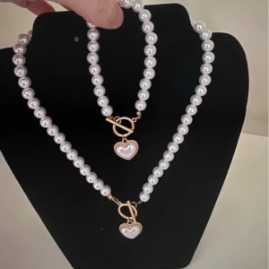 Pärlhalsband och armband med hjärtdetalj - Elegant set med pärlhalsband och matchande armband, båda med vita pärlor och en hjärtformad detalj i guld. Perfekt för att ge en klassisk touch till din stil.