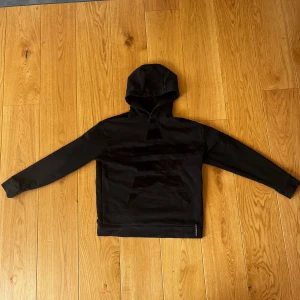 Svart hoodie från Emporio Armani - Snygg svart hoodie från Emporio Armani med ett unikt geometriskt mönster på framsidan. Tröjan har en klassisk huva och långa ärmar, perfekt för en stilren look. Den är tillverkad i ett mjukt material som ger en bekväm passform.
