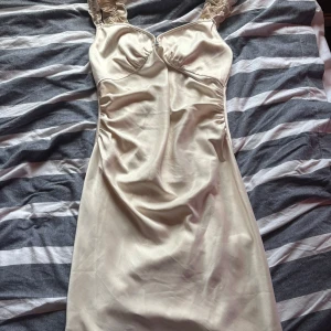 Champagnefärgad klänning från ADOORE - klänning från ADOORE med volangdetaljer på axelbanden. Klänningen har en figurnära passform och är tillverkad i ett glansigt material som ger en lyxig känsla. Perfekt för speciella tillfällen.