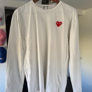 Vit långärmad tröja från Comme des Garçons - Säljer en stilren vit långärmad tröja från Comme des Garçons med det ikoniska röda hjärtat på bröstet. Perfekt för en minimalistisk look. Tröjan är väldigt liten i storleken och skulle säga att den passar s/m bättre.