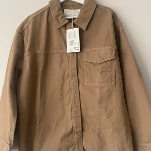 Beige overshirt från COS - Säljer en stilren beige overshirt från COS. Jackan har en klassisk krage och en bröstficka. Perfekt för lager-på-lager-stil och passar till många olika outfits. 🧥