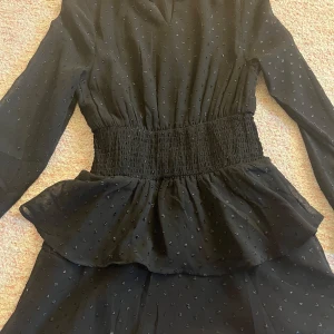 Svart klänning från Gina Tricot - Snygg svart klänning från Gina Tricot med diskret prickigt mönster. Klänningen har långa ärmar och en smockad midja som ger en fin passform. Den har även volangdetaljer vid midjan och en v-ringad hals. Perfekt för en stilren look!