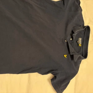 Mörkblå pikétröja från Lyle & Scott 10-11 år - Säljer två mörkblå pikétröjor från Lyle & Scott med klassisk krage och knappslå. De har en broderad gul logotyp på bröstet. Perfekt för en avslappnad men snygg look.