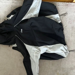 Svart och vit vindjacka från Cortez - Säljer en stilren svart och vit vindjacka från Cortez. Jackan har en justerbar huva och dragkedja framtill. Perfekt för blåsiga dagar och har en sportig look. Passar bra för utomhusaktiviteter.