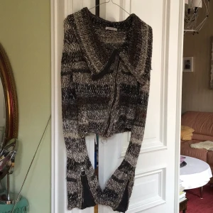 Mönstrad stickad kofta från Sand - Säljer en unik stickad kofta från Sand i jordnära toner av svart, grått och beige. Koftan har en dragkedja framtill och långa ärmar. Perfekt för kyliga dagar!