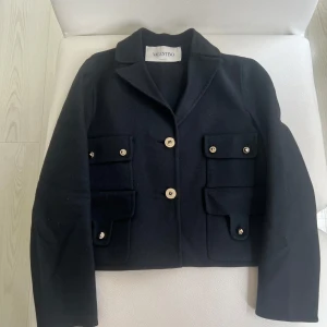 Svart ulljacka från Valentino - Säljer en svart jacka från Valentino i ull med klassisk krage och fyra fickor framtill. Jackan har guldiga knappar och en stilren, tidlös design. Perfekt för dig som vill ha något elegant och exklusivt i garderoben.