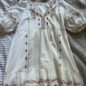 Vit broderad klänning från Odd Molly - Superfin vit klänning från Odd Molly med broderade blommor och volangdetaljer. Klänningen har trekvartsärm och v-ringning med dekorativa sömmar och blommiga broderier längs ärmar och nederkant. Perfekt för sommarens alla tillfällen!