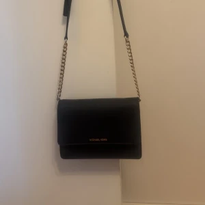 Svart axelväska från Michael Kors - Snygg svart axelväska från Michael Kors med guldfärgad kedja och justerbart axelband. Väskan har flera fack inuti och stängs med lock. Perfekt för att bära det viktigaste med stil.