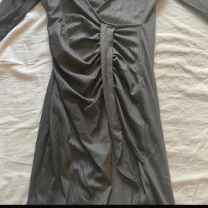 Grå omlottklänning med knyt - Snygg grå omlottklänning med smala ränder och knyt i midjan. Klänningen har v-ringning och ett fint fall. Perfekt för dig som gillar stilrena plagg med en twist.