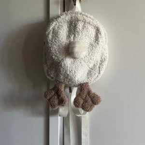 Vit fluffig ryggsäck med ankmotiv - Supergullig vit ryggsäck i teddy-material som ser ut som en anka, komplett med mjuka ben och en liten svans. Justerbara axelband i vitt. Perfekt accessoar för dig som vill sticka ut med något lekfullt och unikt.