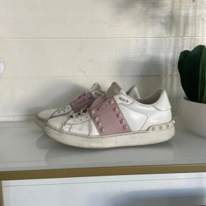 Vita sneakers Valentino Garavani med rosa band - Säljer ett par vita sneakers från Valentino Garavani med breda ljusrosa band över vristen dekorerade med nitar. Skorna har rund tå, platt sula och är tillverkade i skinn. Perfekta för dig som vill ha en stilren men ändå unik look.