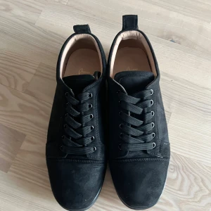 Svarta sneakers från Christian Louboutin - Säljer ett par svarta sneakers från Christian Louboutin med klassisk röd sula och snörning. Skorna är i mocka och har rund tå samt platt sula. Perfekta för dig som vill ha en stilren och lyxig look. Skorna är använda ett fåtal gånger och är nästan som nya (bara skor ingår i köp)