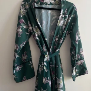 Grön blommig omlottkimono med knytband - Säljer en kort grön kimono/ morgonrock med vackert blommigt mönster i vitt och lila. Klänningen har omlottmodell med v-ringning och knytband i midjan. Långa ärmar och luftig passform, perfekt för sommarens tillfällen. Den är använd för en gång så den är i utmärkt skick. Den är kortare i modellen, ca 77 cm lång. På sista bilden ser ni att den har en liten liten fläck som inte syns alls.🤍🌸💕