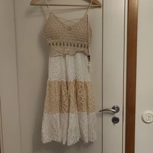 Beige och vit virkad sommarklänning - Säljer en söt klänning med virkad överdel i beige och kjol i lager av spets i beige och vitt. Klänningen har smala axelband och v-ringning med dekorativa band framtill. Perfekt för varma dagar och har en bohemisk känsla.