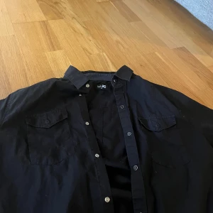 Svart skjorta från JC - Säljer en svart skjorta från JC i storlek M. Skjortan har klassisk krage, knäppning framtill och två bröstfickor med lock. Perfekt för en stilren look och enkel att matcha med olika outfits.