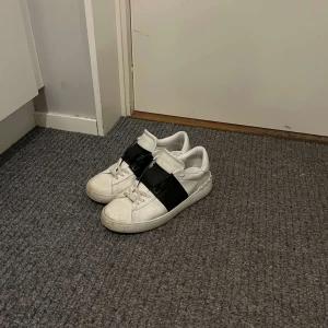 Valentino Open - Säljer ett par vita Valentino Garavani Open sneakers med ett brett svart band över vristen. Skorna har rund tå, platt sula och klassisk snörning. Kommer med originalkartong, dustbag och påse. Perfekta för dig som gillar stilrena och lyxiga sneakers. Skick 8/10. Allt og följer med.