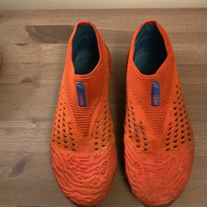 Retro Orange fotbollsskor från Puma - Säljer ett par Retro orangea fotbollsskor från Puma med blå detaljer och mönstrad sula. Skorna har en stickad ovandel med små hål för ventilation och rund tå. Perfekta för dig som vill sticka ut på planen!