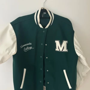 Grön och vit collegejacka från H&M - Snygg grön och vit collegejacka med broderad text 'Minnesota College' och stor M på bröstet. Jackan har vita ärmar, grönt tyg på kroppen, knappar framtill och randiga muddar. Perfekt för en sportig look.