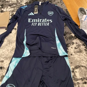Arsenal mörkblå tracksuit - Säljer ett mörkblått Arsenal träningsset från Adidas med ljusblå detaljer. Tröjan har lång ärm och dragkedja vid halsen, samt tryckt klubbmärke och sponsorlogga på bröstet. Byxorna har matchande färg och Adidas-logga.