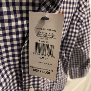 Ny Rutig skjorta från Ralph Lauren - Mörkblå och vit rutig skjorta från Ralph Lauren i bomullspoplin. Klassisk krage och knäppning framtill, med liten orange broderad logga på bröstet. Perfekt för en stilren look.