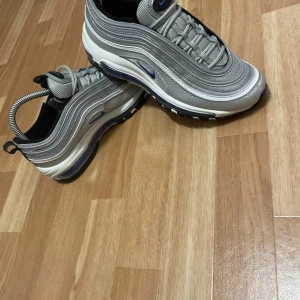 Nike Air Max 97 Silver Bullet - ljer dessa grymt snygga Nike Air Max 97 i silver med stilrena blå detaljer – en riktig klassiker som aldrig går ur tiden! De här skorna kombinerar futuristisk design med den bekväma Air-dämpningen Nike är kända för. Perfekta både till vardags och när du vill lyfta din outfit ett snäpp!  Skorna är i superbra skick – nästan som nya – och redo för en ny ägare som vill sticka ut med stil.  Detaljer: 	•	Modell: Nike Air Max 97 	•	Färg: Silver/Blå (riktigt clean färgkombo!) 	•	Storlek: [38,5] 