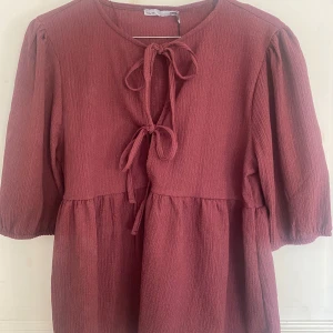 Vinröd blus med knytning och puffärmar - Söt röd blus med knyt framtill och puffiga ärmar. Blusen har en avslappnad passform och är tillverkad i ett strukturerat tyg som ger en mjuk känsla. Perfekt för vår och sommar!