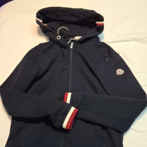 Mörkblå hoodiejacka från Moncler - Snygg mörkblå hoodiejacka från Moncler med dragkedja och huva. Jackan har Moncler-logga på bröstet och rödvita detaljer vid ärmslut och huva. Perfekt för en sportig och avslappnad stil.möts i nrkp