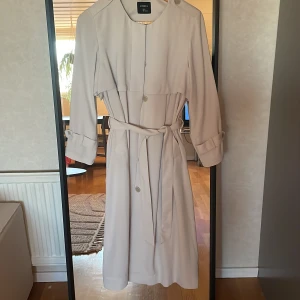 Beige trenchcoat från Lindex - Stilren ljusbeige trenchcoat från Lindex med knytskärp i midjan och knappar framtill. Jackan har långa ärmar med spänndetaljer vid ärmsluten och klassisk krage. Perfekt för övergångssäsonger och enkel att matcha till olika outfits.