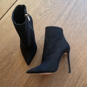 Svarta mocka stövletter med klack - Säljer ett par svarta stövletter i mocka med hög smal klack och spetsig tå. Skorna har dragkedja på sidan för enkel på- och avtagning. Perfekta för dig som vill ha en stilren och elegant look.
