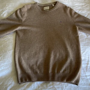 Beige kashmirtröja från GANT - Säljer en beige kashmirtröja från GANT med rund hals och diskret broderad logga på bröstet. Tröjan har ribbade muddar vid ärmslut och nederkant. Perfekt för dig som vill ha en klassisk och stilren look.