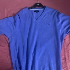 Blå v-ringad tröja från GANT - Säljer en blå v-ringad tröja från GANT med diskret broderad logga på bröstet. Tröjan har lång ärm och är tillverkad i mjukt material, perfekt för lager-på-lager eller som den är.