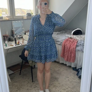 Blå mönstrad omlottklänning - Säljer en blå kortklänning med vitt mönster och omlottliv. Klänningen har långärmade, lätt transparenta ärmar och markerad midja. Kjolen är volangig och tyget är tunt och luftigt, perfekt för varmare dagar.