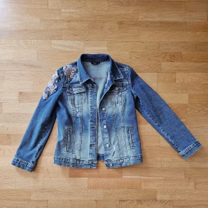 Jeansjacka med blommig brodyr - Snygg blå jeansjacka med blommig brodyr på ena axeln. Klassisk modell med knappar framtill och två bröstfickor. Jackan har lång ärm och en normal passform. Perfekt för att lyfta din outfit med en unik detalj. Som en ny jacka!