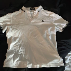 Vit pikétröja från Fred Perry - Snygg vit pikétröja från Fred Perry med korta ärmar och klassisk krage. Tröjan har en broderad logga på bröstet och svarta detaljer vid ärmslut och krage. Perfekt för en stilren look!